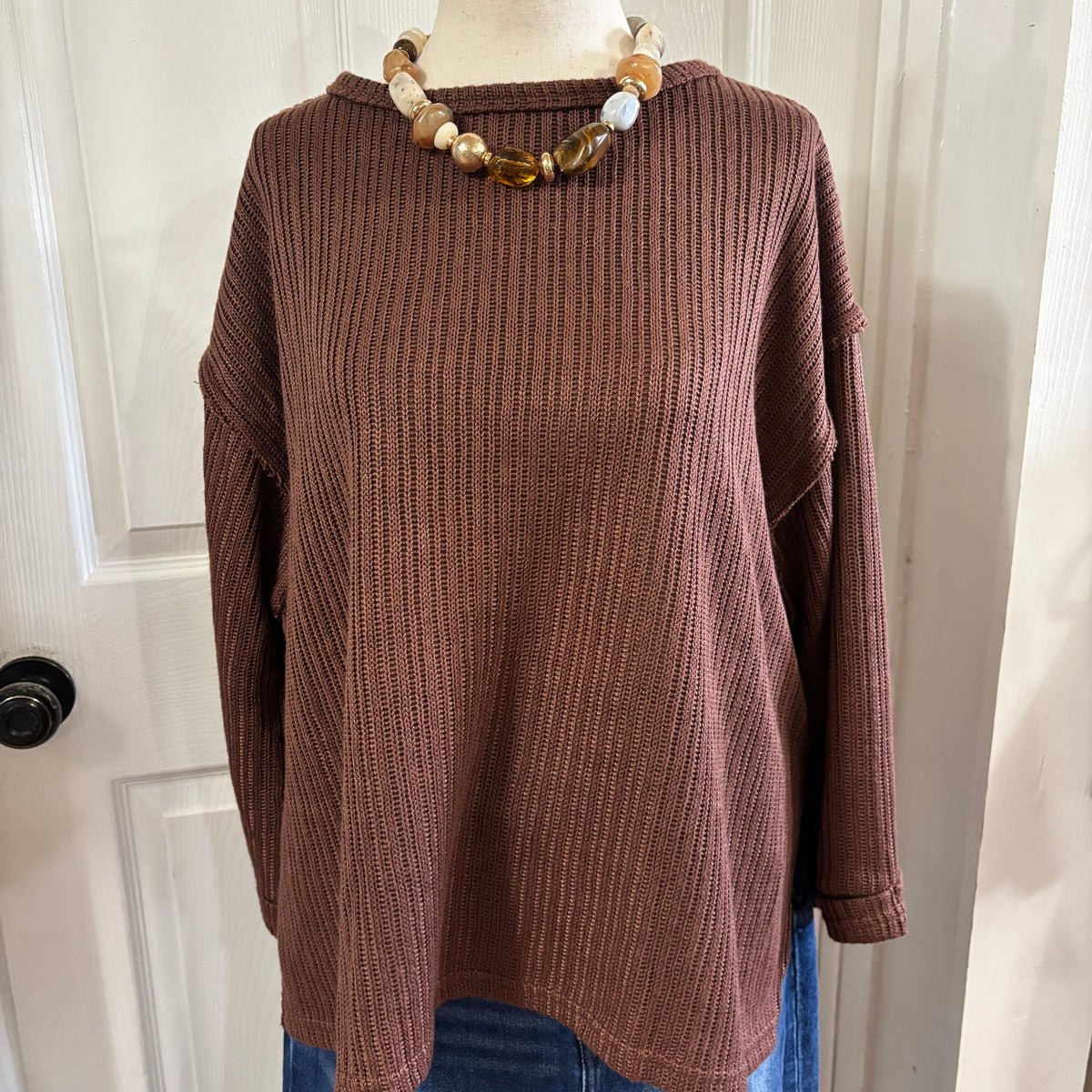 A brown ribbed knit long-sleeve sweater displayed on a mannequin torso, styled with a chunky beaded necklace and blue jeans, set against a white paneled door background.