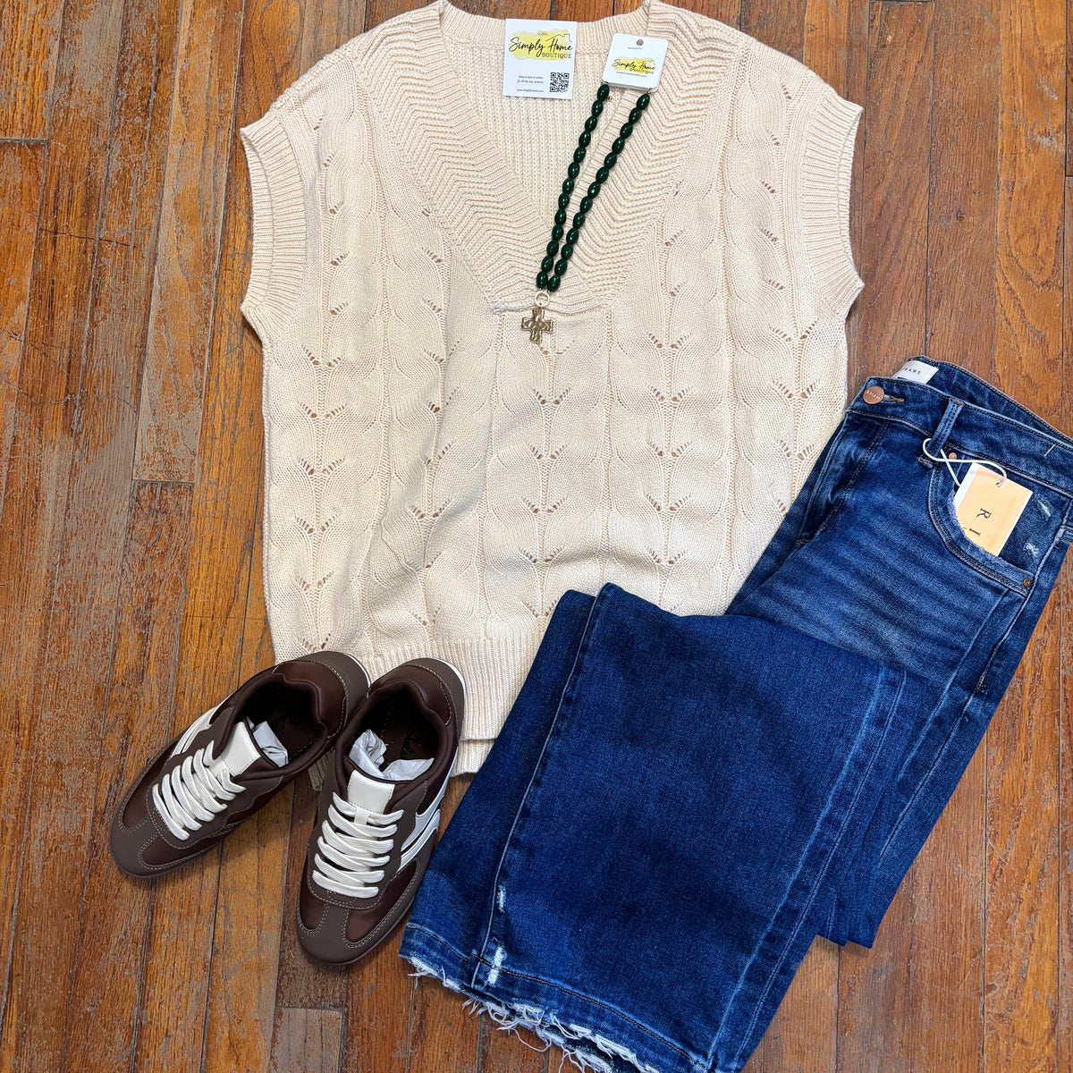 A styled flat-lay outfit on a wooden floor featuring a cream cable-knit sweater vest, dark blue denim jeans, brown sneakers, and a dark green beaded necklace.