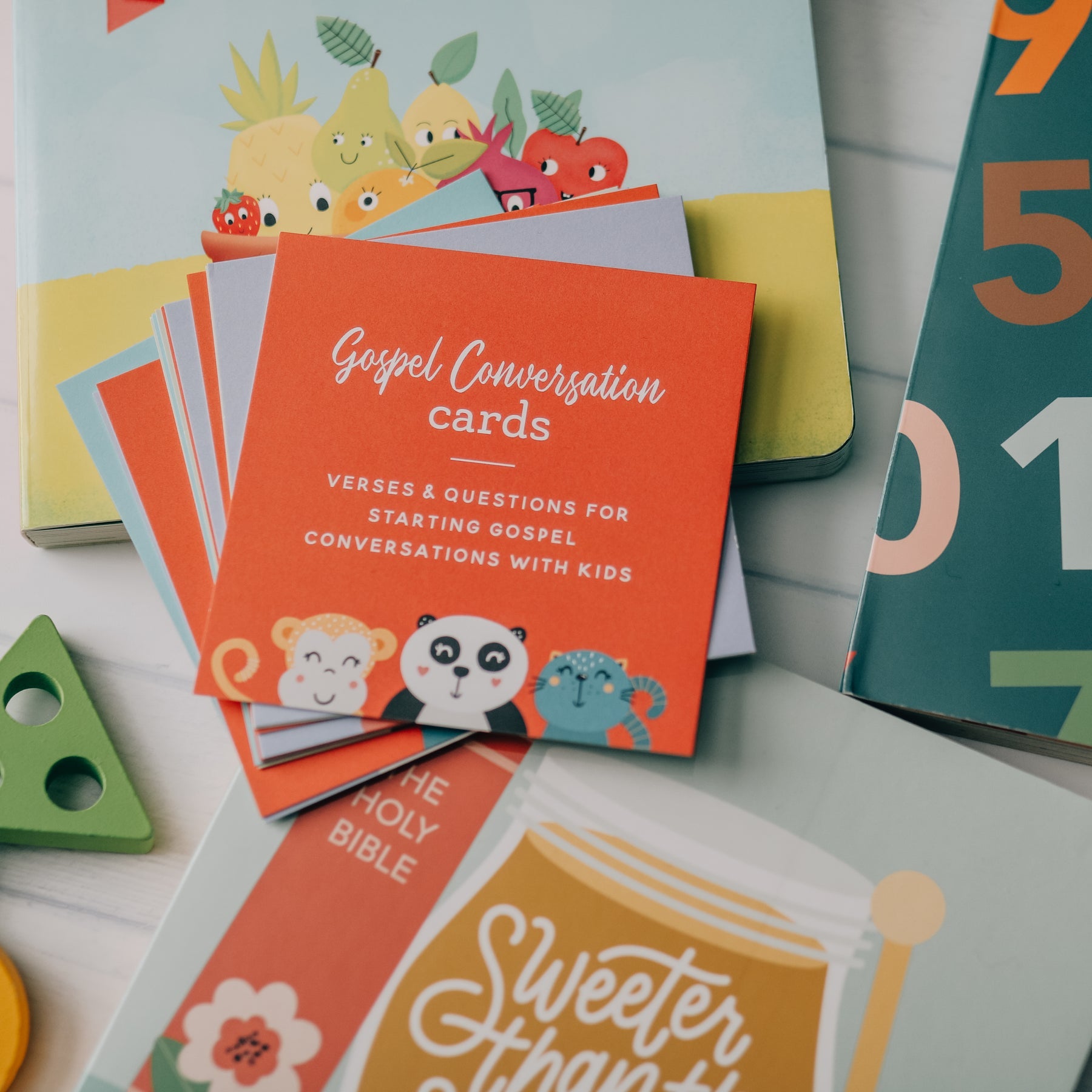 A stack of colorful Gospel Conversation cards for kids, featuring a bright coral-orange cover with white text and cartoon animal illustrations, displayed on a light surface with other children's books and toys nearby.
