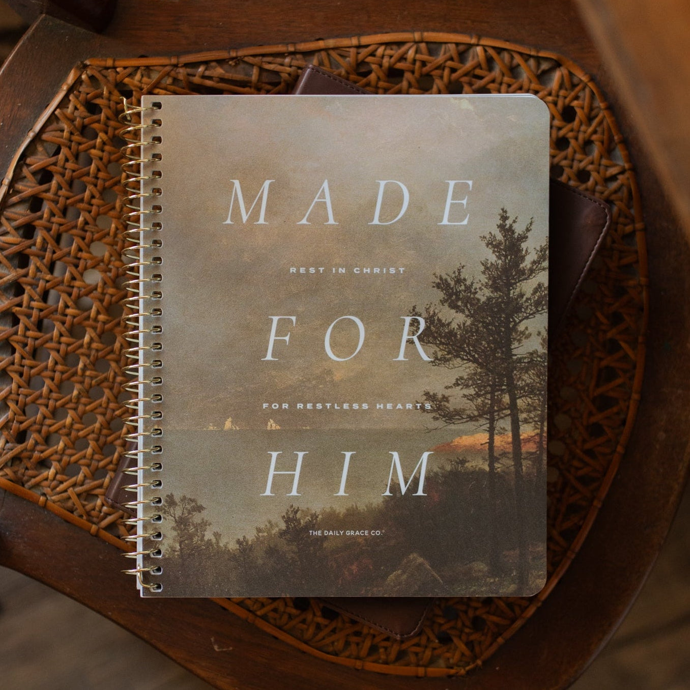 A spiral-bound notebook with a scenic cover reading 'MADE FOR HIM' lies on a woven brown tray over a wooden surface.