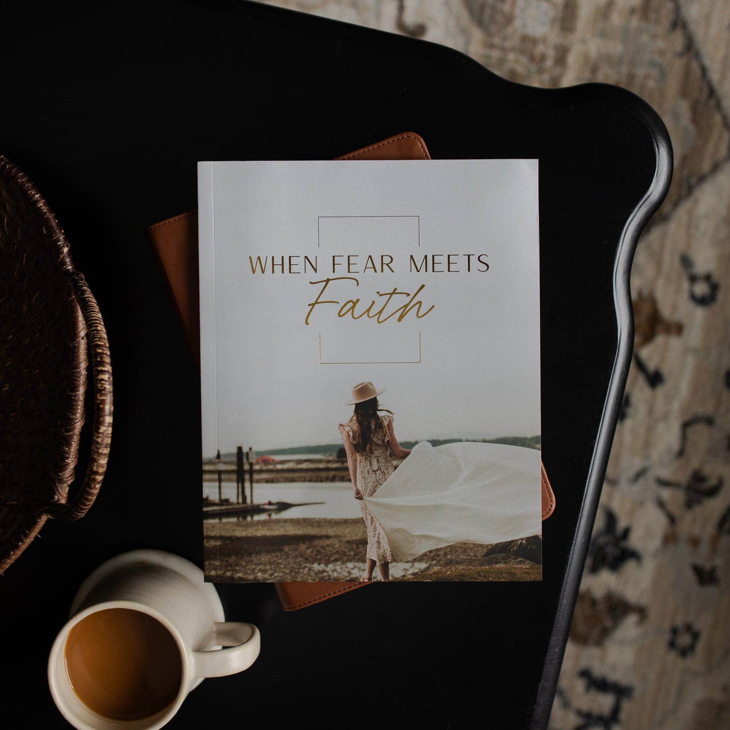 A printed book or journal titled 'When Fear Meets Faith' lies on a black tray, with a coffee cup nearby and a patterned rug in the background. The cover shows a woman standing outdoors with a flowing white dress.